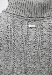 Pull à col roulé gris en maille avec des motifs torsadés et une petite étiquette métallique portant la marque Barbour au dos.