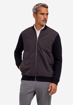 Man with short gray hair wearing a black zip-up jacket with knit sleeves over a white shirt and gray pants, looking to the side.