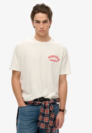 T-shirt en coton blanc avec un graphisme rouge "Superdry Los Angeles", associé à un jean bleu et une chemise à carreaux nouée autour de la taille.
