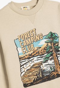 Sudadera beige con cuello de canalé que presenta un estampado gráfico de una escena de bosque, con "FOREST CAMPING CLUB" y un árbol a la izquierda.