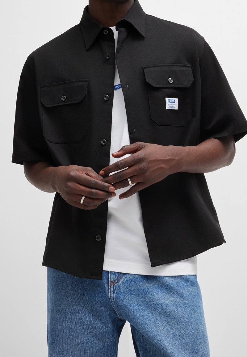 Zwart shirt met korte mouwen, twee borstzakken, knoopsluiting en een merklabel. Draagt over een wit T-shirt en blauwe spijkerbroek.