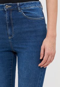 Denim jeans in dark blue with a smooth texture, featuring five pockets, light stitching, and a zip button fly closure.