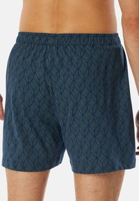 Bañador azul marino con un patrón geométrico de hojas; cintura elástica y corte relajado; tejido suave con una textura lisa.