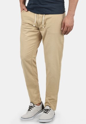 Man wearing beige drawstring trousers, navy shirt, and white sneakers with black laces standing against plain background.