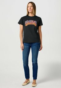 Camiseta de algodón negra con un gráfico colorido de "Wrangler" en la parte frontal, combinada con jeans azules y zapatos planos beige. Diseño simple y estilo casual.