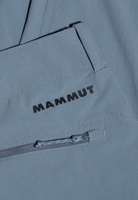 Vaalea sininen kangas, jossa on sileä pinta, varustettuna vetoketjullisella taskulla ja mustalla "MAMMUT"-logolla edessä. Yksinkertainen, toiminnallinen muotoilu.