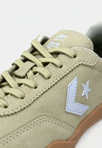 Beige ruskindsneaker med beige snørebånd, hvid stjerne- og pil-logo på siden samt brun sål, der viser detaljerede syninger og tekstur.