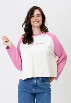 Jonge vrouw glimlacht, draagt een wit-roze longsleeve met de tekst "Lucky Girls Club" en een blauwe spijkerbroek met met studs versierde zakken.