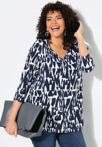 V-Ausschnitt-Top mit einem navyblauen und weißen abstrakten Muster, ¾-Ärmel, kombiniert mit einer grauen Clutch. Material aus Baumwollmischung.