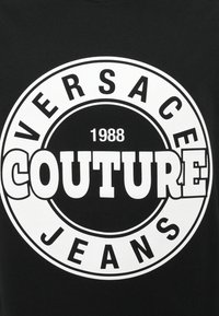 Juodas medvilnės marškinėlis su dideliu apvaliu grafiku, kuriame didelėmis baltomis raidėmis parašyta "VERSACE" ir "COUTURE JEANS", su 1988 metų numeriu.