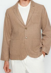 Giacca in misto lana beige con revers a punta, due tasche frontali e chiusura a due bottoni. Texture liscia, vestibilità leggermente oversize.