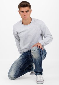 Felpa a girocollo grigia chiara in cotone abbinata a jeans blu in denim strappati. Il modello è in ginocchio, mostrando il tessuto consumato e i colori a contrasto.