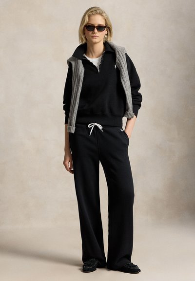 Giovane donna che indossa occhiali da sole neri, felpa nera con zip, pantaloni larghi neri, maglione grigio sulle spalle e scarpe nere.