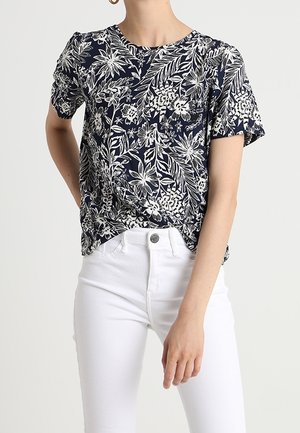 Vrouw draagt een marineblauwe top met korte mouwen en witte bloemenprint, ingestopt in hoge witte broek.