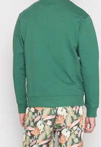 Sudadera verde hecha de un tejido suave, con un diseño simple y puños y dobladillo acanalados. Combinada con pantalones de estampado floral.