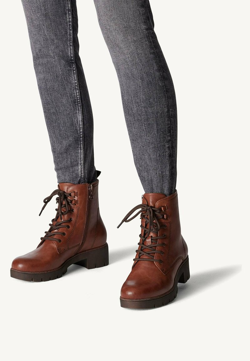 Botas de cuero marrón hasta el tobillo con puntera redondeada, frente con cordones y cremallera lateral. Tacón grueso y suela de goma texturizada. Llevadas con jeans grises.