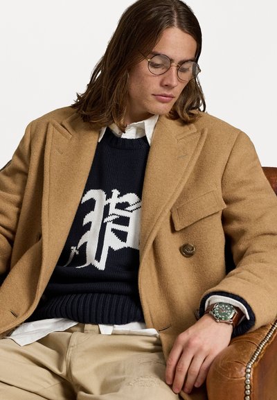 Ung man med axellångt brunt hår som bär runda glasögon, en kamelfärgad kappa, en marinblå tröja med vit design, beige byxor och en armbandsur.