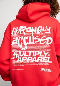 Personne aux cheveux bouclés portant un sweat à capuche rouge avec un grand texte blanc "WRONGLY ACCUSED" et "MULTIPLY APPAREL" au dos.