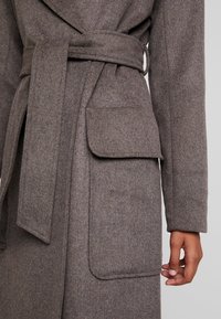 Brauner Wollmantel mit Wickeldesign, großen Seitentaschen und passendem Gürtel. Die Textur ist weich und leicht fusselig.