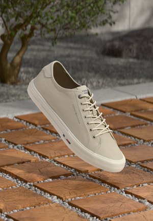 Beige cotton canvas sneaker with flat laces, rounded toe, and rubber sole, featuring a small logo and colored accent on the side.
