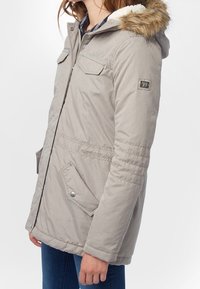 Světlá šedá parka s kapucí s kožešinovou podšívkou, zipem v přední části, dvěma náprsními kapsami a bočními kapsami; prošívané detaily na rukávech a lemu.