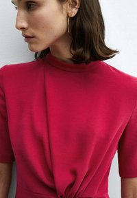 Úzky šaty z živého ružovo-červeného materiálu s vysokým výstrihom, krátkymi rukávmi a uzlovým detailom v oblasti pása, ktorý vytvára hladký siluetu.