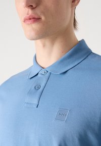 Svetlomodrá polo košeľa s jemnou textúrou, krátkymi rukávmi a zapínaním na dva gombíky. Obsahuje malé vyšívané logo značky.