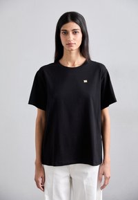 Black short-sleeve t-shirt made of cotton, featuring a round neckline and a small gold logo accent on the chest. Simple, straight-cut design.