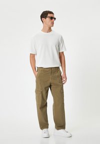 Khaki cargo-housut, joissa on useita sivutaskuja, yhdistettynä valkoiseen ribattuun lyhythihaiseen paitaan ja valkoisiin lenkkareihin. Mallilla on tummat aurinkolasit.
