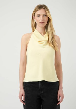 Femme blonde portant un haut sans manches jaune pâle à col bénitier et un pantalon noir, debout devant un fond clair et uni.