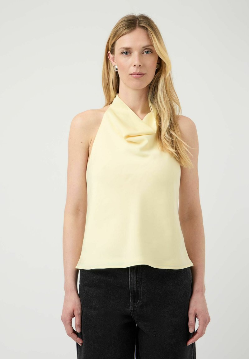 Femme blonde portant un haut sans manches jaune pâle à col bénitier et un pantalon noir, debout devant un fond clair et uni.