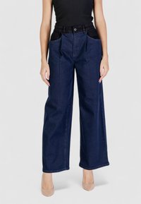 Jeans in denim scuro a gamba larga con ampie tasche anteriori nere, taglio dritto e tessuto non elasticizzato. La cintura è dotata di passanti per la cintura.