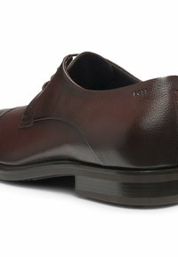 Scarpe eleganti in pelle marrone con una texture liscia, punta arrotondata e un sottile dettaglio di branding. Presentano una suola bassa in gomma nera.