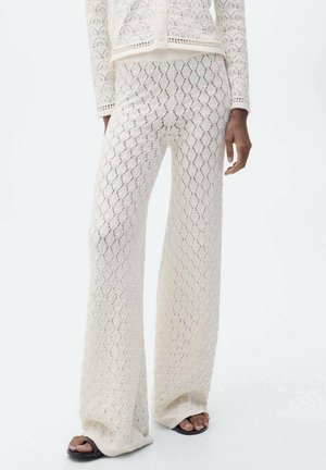 Weiße Spitzenstrickhose mit weitem Bein, getragen mit passendem, geknöpftem Cardigan und schwarzen Sandalen mit offenem Zehenbereich, dargestellt von der Taille bis zu den Füßen.