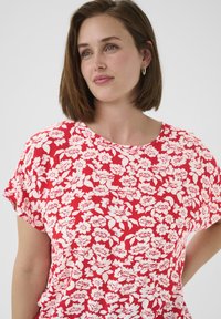 Blouse florale rouge avec des motifs de fleurs blanches, manches courtes, encolure ronde et texture de tissu douce. Convient pour une tenue décontractée.