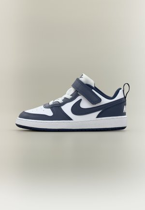 Adidaș alb și bleumarin Nike cu baretă Velcro, șireturi, logo negru Swoosh și buclă de prindere pe fundal gri deschis.