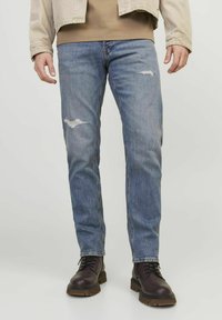 Vaqueros de mezclilla azul claro con corte recto, que presentan detalles desgastados y un lavado medio. Combinados con botas de cuero marrón oscuro con cordones.