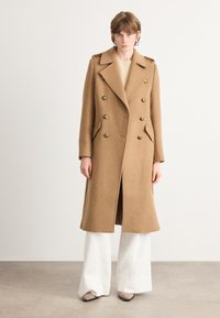 Palton din lână bej, cu două rânduri de nasturi mari, guler cu colțuri și buzunare laterale, purtat cu pantaloni largi albi și un top neutră.