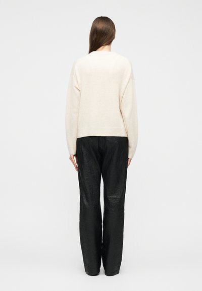 Γυναίκα με μακριά καστανά μαλλιά, που φοράει ένα κρεμ πουλόβερ και χαλαρό μαύρο παντελόνι με υφή, στέκεται μπροστά σε μια απλή λευκή επιφάνεια.