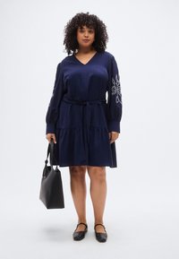 Φόρεμα σε ναυτικό μπλε, μήκους έως το γόνατο, με V-λαιμόκοψη, μακριά μανίκια με λευκή κεντητή ανθοδέσμη και ζώνη στη μέση. Συνδυάζεται με μαύρη τσάντα χειρός και επίπεδα παπούτσια.