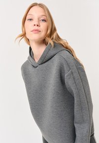 Felpa grigia con cappuccio, dal tocco morbido, caratterizzata da una vestibilità comoda e maniche leggermente allungate, su uno sfondo semplice.