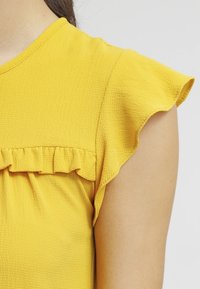 Top giallo con maniche corte a volant e tessuto texture. Presenta un collo rotondo e un dettaglio arricciato sotto il volant all'altezza del busto.