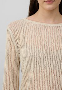 Sweter z dzianiny w kremowym kolorze z otwartym, teksturowanym wzorem. Posiada szeroką, łodziastą dekoltę i długie rękawy, eksponując lekką konstrukcję.