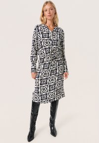 Mustavalkoinen kuviollinen mekko, jossa on pitkät hihat, V-pääntie ja kiristettävä vyötärö. Yhdistetty kiiltäväksi mustien polvipitkien saappaiden kanssa.