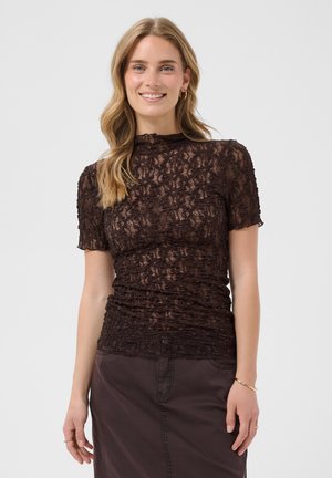 Femme souriante, portant un haut en dentelle brune transparente à manches courtes et une jupe brune assortie, avec des cheveux ondulés et des créoles dorées.