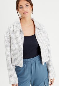 Weiße Tweedjacke mit blauem und elfenbeinfarbenem Karomuster, kurzes Design, breiter Kragen und verdeckter Reißverschluss vorne, kombiniert mit einem schwarzen Oberteil und blauen Hosen.