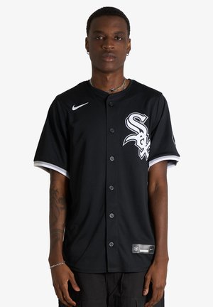 Giovane uomo che indossa una maglia da baseball nera dei Chicago White Sox con logo e bottoni bianchi, in piedi davanti a uno sfondo bianco neutro.