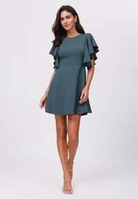 Teal kort klänning med fjärilsärmar, figursydd bodice och utsvängd kjol. Slät tygl och rena linjer. Bärs med nude remsandaler.