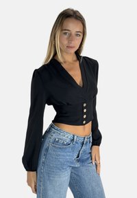 Zwarte wikkelblouse met lange, geplooide mouwen, voorzien van een V-hals en drie knoopaccenten bij de taille, gecombineerd met lichtblauwe spijkerbroek.