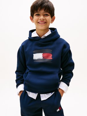 Navyblauwe hoodie van zacht materiaal met een vooraanstaand grafisch ontwerp van witte en rode penseelstreken. Gelaagd over een witte blouse. Soepele textuur.